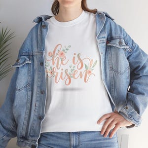 Könnte beinhalten: Weißes T-Shirt mit dem Schriftzug "He is Risen" in Schreibschrift, verziert mit pfirsichfarbenen Rosen und grünen Blättern. Das Shirt wird mit einer hellblauen Jeansjacke und blauen Jeans getragen.