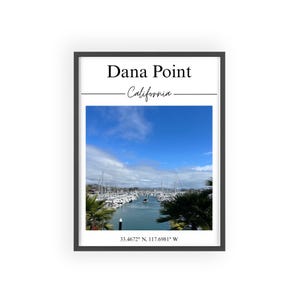 Könnte beinhalten: Gerahmter Druck von Dana Point, Kalifornien, mit einem Yachthafen voller Boote unter blauem Himmel. Das Bild enthält den Text "Dana Point - California" oben. Palmen sind auf beiden Seiten des Hafens zu sehen, mit den Koordinaten 33.4672° N, 117.6981° W unten.