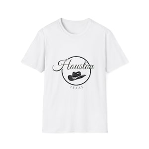 Könnte beinhalten: Weißes T-Shirt mit einem schwarzen kreisförmigen Design. Das Design enthält das Wort "Houston" in Schreibschrift, eine Cowboyhut-Illustration und das Wort "TEXAS". Das T-Shirt besteht aus einem weichen, leichten Material.