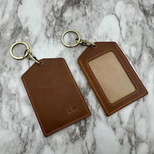 May include: Two brown leather keychains with brass-colored hardware. One keychain has a clear window, and the other has the initials "BM" embossed on the front. Both have a rectangular shape with rounded edges.