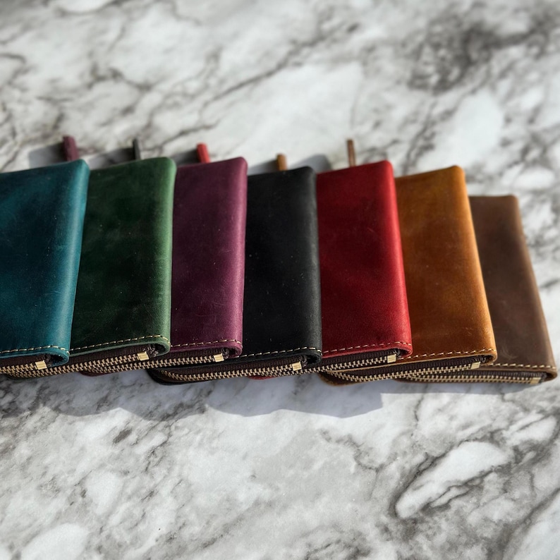 Puede incluir: Una colecci&oacute;n de siete carteras de cuero en varios colores, incluyendo turquesa, verde, burdeos, negro, rojo, marr&oacute;n claro y marr&oacute;n. Cada cartera tiene una cremallera dorada y una leng&uuml;eta de cuero a juego.