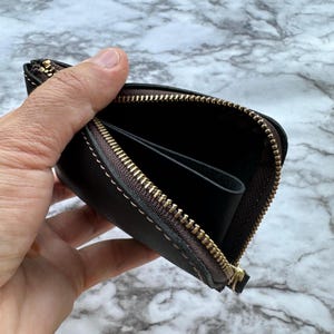 May include: A dark brown leather wallet with a gold zipper is held open, revealing the interior. The wallet has a stitched border and appears to be empty. The background is a marble pattern.