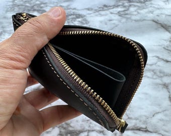 May include: A dark brown leather wallet with a gold zipper is held open, revealing the interior. The wallet has a stitched border and a small pocket inside. The zipper is fully open, showing the inside of the wallet.