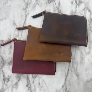 Puede incluir: Tres monederos de cuero con cremallera en tonos burdeos, marr&oacute;n y beige. Cada monedero tiene una cremallera dorada y una peque&ntilde;a leng&uuml;eta de cuero. Los monederos est&aacute;n grabados con iniciales o un nombre. Los monederos son rectangulares y parecen hechos a mano.