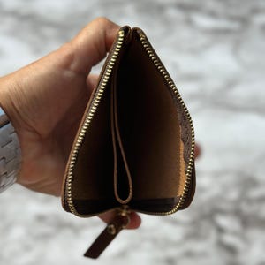 Puede incluir: Cartera de cuero marr&oacute;n abierta con cremallera dorada. La cartera se sostiene abierta, revelando los compartimentos interiores y una peque&ntilde;a leng&uuml;eta de cuero unida a la cremallera.
