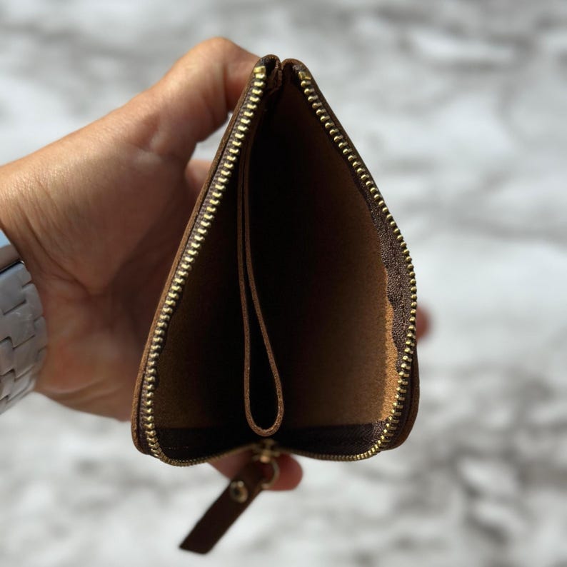 Puede incluir: Cartera de cuero marr&oacute;n abierta con cremallera dorada. La cartera se sostiene abierta, revelando los compartimentos interiores y una peque&ntilde;a leng&uuml;eta de cuero unida a la cremallera.