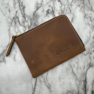 Puede incluir: Un monedero de cuero marr&oacute;n con cremallera dorada y el texto "EDWARD R" en relieve. El monedero rectangular tiene un borde cosido. El cuero tiene un aspecto envejecido.