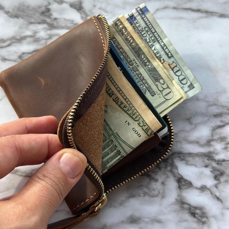 Puede incluir: Una cartera de cuero marr&oacute;n con cierre de cremallera, parcialmente abierta para revelar varios billetes de d&oacute;lar estadounidense y tarjetas de cr&eacute;dito. La cartera contiene varias denominaciones.