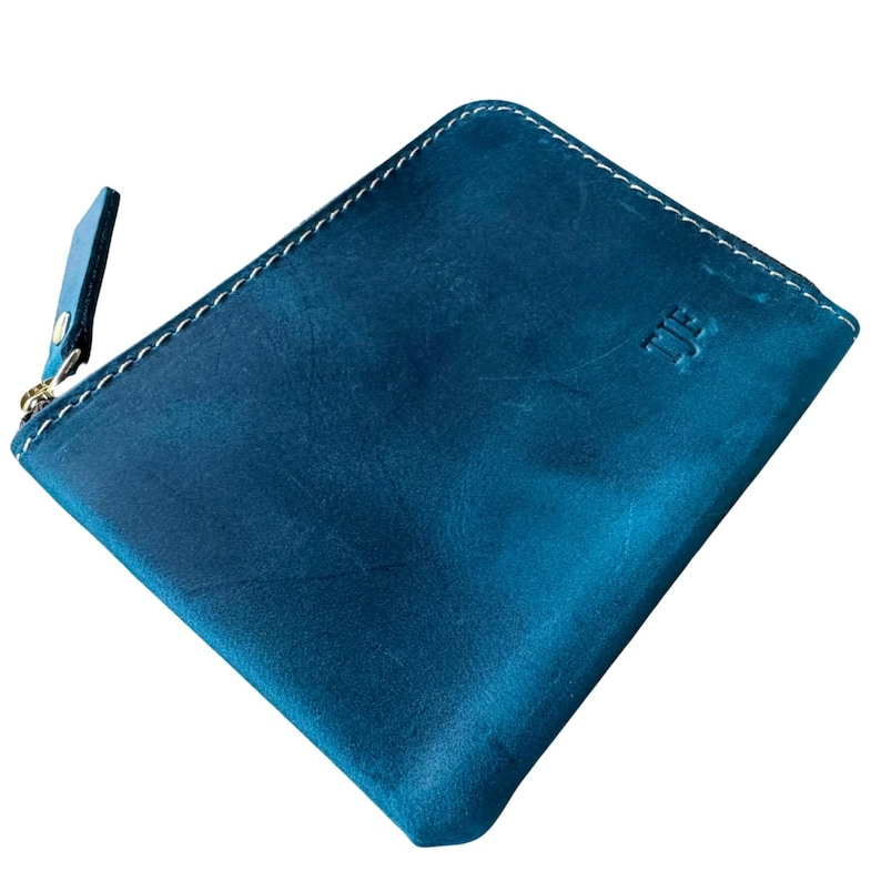 Puede incluir: Una cartera de cuero color turquesa con cierre de cremallera y costuras blancas. La cartera tiene una peque&ntilde;a leng&uuml;eta de cuero en la cremallera y un logotipo en relieve en la parte delantera. La cartera es de forma cuadrada.