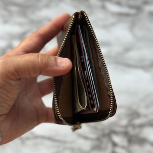 Puede incluir: Un monedero de cuero marr&oacute;n con cremallera dorada est&aacute; parcialmente abierto, revelando varias tarjetas de cr&eacute;dito y billetes doblados. El monedero se sostiene en una mano, mostrando su dise&ntilde;o compacto y capacidad de almacenamiento.