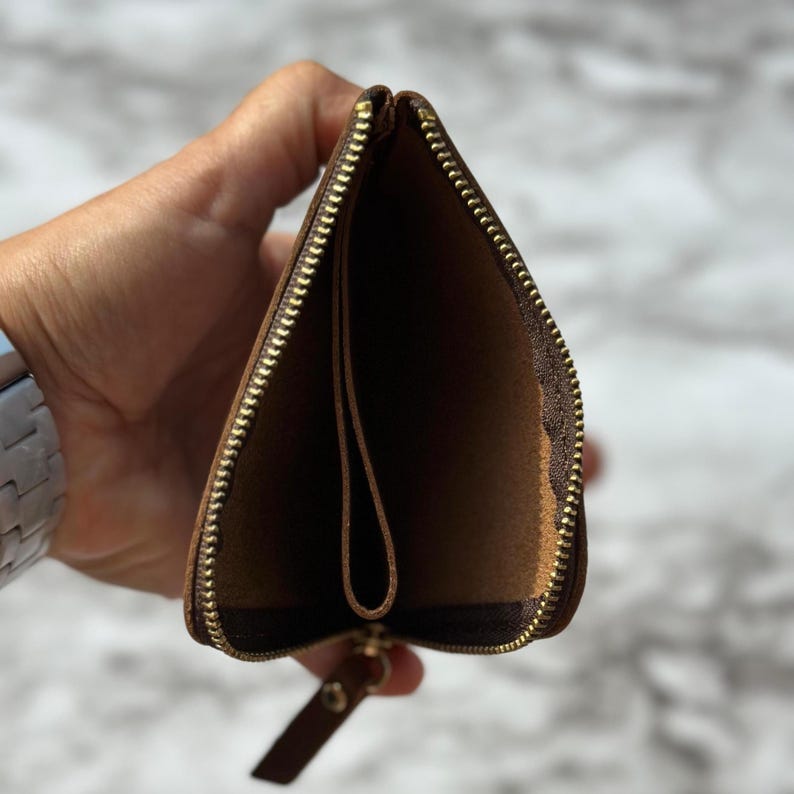 Puede incluir: Cartera de cuero marr&oacute;n abierta con cremallera dorada. El interior es de un tono marr&oacute;n m&aacute;s claro. La cartera se sostiene abierta, revelando el interior. El tirador de la cremallera tambi&eacute;n es de cuero marr&oacute;n.