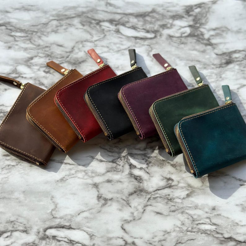 Puede incluir: Colecci&oacute;n de siete carteras de cuero en varios colores, incluyendo marr&oacute;n, rojo, negro, morado, verde y turquesa. Cada cartera tiene una cremallera dorada y una peque&ntilde;a leng&uuml;eta de cuero. Disposici&oacute;n diagonal.