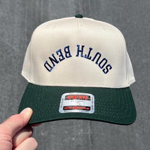 Op de afbeelding: Een crème en bosgroene baseballcap met de tekst "SOUTH BEND" geborduurd in marineblauw. De cap heeft een gebogen klep en een kleine groene knoop bovenop. Het merklabel "OTTO" is zichtbaar op de klep.