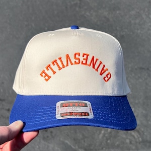 May include: A white and blue baseball cap with the orange word "GAINESVILLE" arched across the front. The cap has a blue brim and a blue button on top. The cap has a tag that says "OTTO".