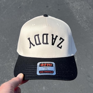 Pode incluir: Boné de beisebol creme e preto com a palavra "ZADDY" bordada em preto. O boné tem uma aba preta e um botão preto na parte superior. A etiqueta diz "OTTO CLASSIC FIT OSFM MID PROFILE MAXIMUM COMFORT".