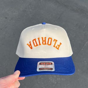 May include: A white and blue baseball cap with the word "FLORIDA" embroidered in orange. The cap has a curved brim and a blue button on top. The brand tag is visible on the front.