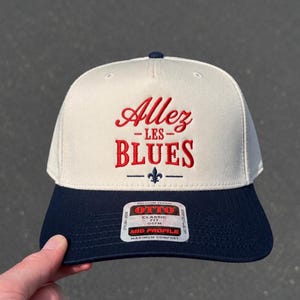 May include: A cream-colored baseball cap with a navy blue brim and a top button. The front features red embroidered text that reads Allez LES BLUES, with a small navy blue fleur-de-lis symbol below. A sticker on the brim reads OTTO CLASSIC FIT.