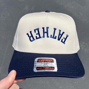 May include: A cream-colored baseball cap with a navy blue brim and the word "FATHER" embroidered in navy blue. The cap has a classic fit and a mid-profile design.
