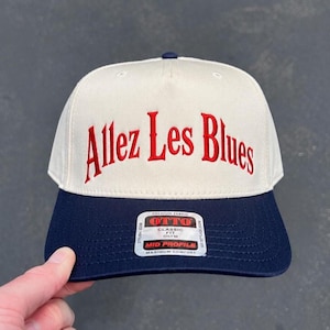 May include: A two-tone baseball cap with an off-white crown and a navy blue brim. The front features the embroidered red text Allez Les Blues. A rectangular sticker on the brim displays the text OTTO PREMIUM FABRIC CLASSIC FIT OSFM MID PROFILE.