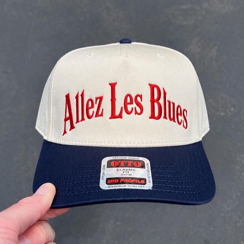 May include: A two-tone baseball cap with an off-white crown and a navy blue brim. The front features the embroidered red text Allez Les Blues. A rectangular sticker on the brim displays the text OTTO PREMIUM FABRIC CLASSIC FIT OSFM MID PROFILE.