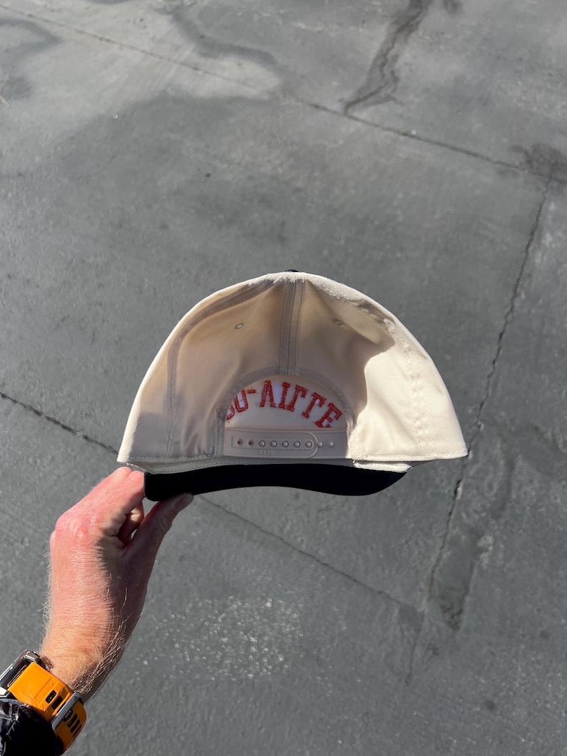 May include: A cream-colored baseball cap with a black brim and a plastic snapback closure. The interior back panel features the red embroidered text "LAFITTE" in a bold, arched font, positioned directly above the adjustable snapback strap.