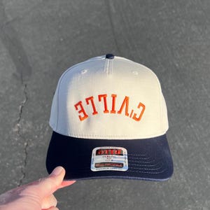 Puede incluir: Una gorra de béisbol de dos tonos con una corona de color beige claro y una visera de color azul marino oscuro. El frente presenta la palabra C'VILLE bordada en hilo naranja en una orientación arqueada e invertida. Una etiqueta en la visera.