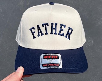 May include: A baseball cap with a cream-colored crown and a navy blue brim. The word "FATHER" is embroidered in navy blue across the front. The cap has a curved brim and a button on top. A tag with the brand name is visible.