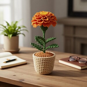 May include: A crocheted marigold flower in a beige pot. The flower has orange petals and a green stem and leaves. The pot is made of beige yarn. The flower is sitting on a wooden table.