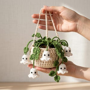 Puede incluir: Una maceta colgante de ganchillo en color beige claro, con enredaderas y hojas verdes tejidas. Varias figuras pequeñas de ganchillo en forma de fantasma blanco, con ojos de cuentas negras, están unidas a las enredaderas y cuelgan de la cesta.