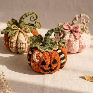 Puede incluir: Tres calabazas de ganchillo con tallos y hojas detallados. Una es beige con un patrón de cuadros, una es naranja con una cara de jack-o'-lantern, una luna creciente y estrellas, y una es rosa claro con un lazo y un pequeño corazón.