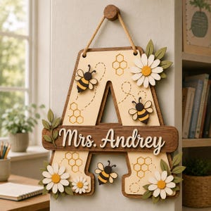 以下が含まれることがあります： Mrs. Andreyという白い筆記体の文字が入った、木製のAの形をした壁掛けサイン。立体的なハチ、黄色い中心を持つ白いデイジー、緑の葉、そして刻印されたハニカム模様で装飾されており、ロープで吊り下げられています。