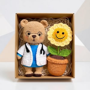 May include: A crocheted teddy bear in a doctor's coat and spectacles, beside a crocheted sunflower in a pot, presented in a cardboard box with paper filler. The bear is tan, the coat is white, and the flower is yellow.