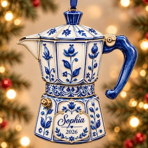 May include: A ceramic moka pot ornament featuring a white base with blue floral patterns, a blue handle, and a blue lid. The front displays a decorative label with the name Sophia and the year 2026 printed in blue script and block lettering.