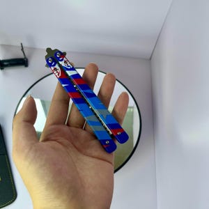 K&ouml;nnte beinhalten: Ein dekoratives Butterflymesser mit blauem, roten und wei&szlig;en Design. Das Messer hat eine silberne Klinge und wird in einer Hand gehalten. Der Griff hat ein Streifenmuster und eine Grafik.