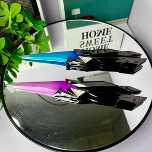 Puede incluir: Dos abridores de cartas decorativos con un diseño futurista. Uno tiene una hoja azul, el otro una hoja morada. Ambos tienen mangos negros con detalles plateados. Los abridores de cartas se exhiben sobre una superficie reflectante.