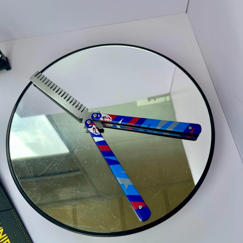 K&ouml;nnte beinhalten: Ein Butterflymesser mit silberner Klinge und einem Griff mit blauem, roten und wei&szlig;en geometrischen Muster. Das Messer ist ge&ouml;ffnet und zeigt die kammartige Klinge. Der Griff hat ein kreisf&ouml;rmiges Design mit einem Logo.