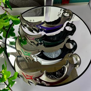 May include: A collection of five curved, silver-toned karambit knives with various accent colors, including purple, green, red, and black. The knives are displayed on a reflective surface, showcasing their intricate designs and sharp edges. The blades have a curved shape with a finger ring.