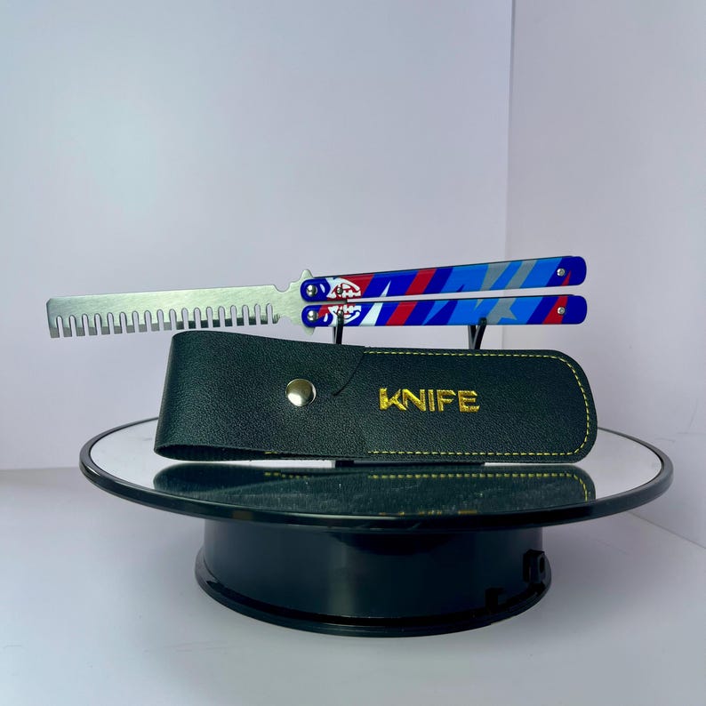 K&ouml;nnte beinhalten: Ein Butterflymesser mit silberner Klinge und blauen, roten und wei&szlig;en Griffen. Das Messer ist ge&ouml;ffnet und zeigt die gezahnte Kante. Eine schwarze Lederscheide mit dem Wort "KNIFE" in goldfarbenen Buchstaben befindet sich darunter.