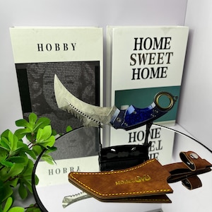 May include: A curved karambit knife with a patterned blade and blue handle is displayed on a stand. The knife is accompanied by a brown leather sheath. The background includes two books with the words "HOBBY" and "HOME SWEET HOME".