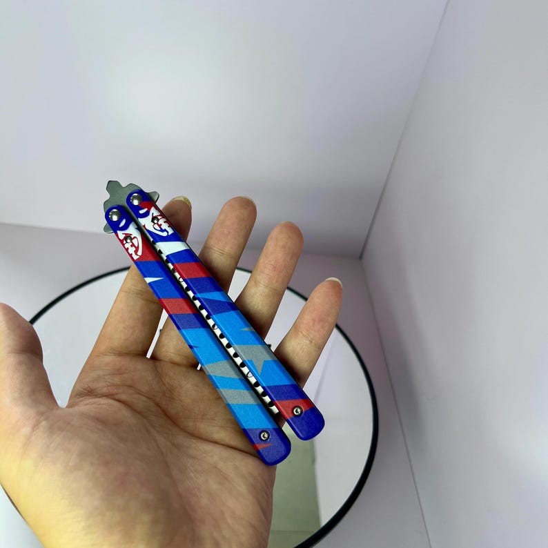 K&ouml;nnte beinhalten: Ein Butterflymesser mit blauem, rotem und grauem Design. Die Griffe weisen ein sich wiederholendes Muster aus Streifen und einer Cartoonfigur auf. Die Klinge ist silberfarben. Das Messer wird in einer Hand gehalten.