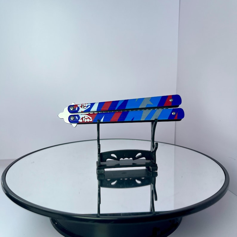 K&ouml;nnte beinhalten: Ein Butterflymesser mit einem geometrischen Muster in Blau, Rot und Grau. Das Messer wird auf einem schwarzen St&auml;nder vor wei&szlig;em Hintergrund pr&auml;sentiert. Die Klinge hat ein Design mit roten und wei&szlig;en Akzenten.