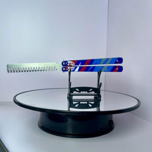K&ouml;nnte beinhalten: Ein Butterflymesser mit silberner Klinge und blau-rot-wei&szlig;em Griffdesign. Der Griff weist ein geometrisches Muster und ein Logo auf. Das Messer wird auf einem schwarzen St&auml;nder mit verspiegelter Oberfl&auml;che pr&auml;sentiert.