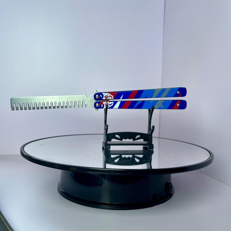 K&ouml;nnte beinhalten: Ein Butterflymesser mit silberner Klinge und blau-rot-wei&szlig;em Griffdesign. Der Griff weist ein geometrisches Muster und ein Logo auf. Das Messer wird auf einem schwarzen St&auml;nder mit verspiegelter Oberfl&auml;che pr&auml;sentiert.