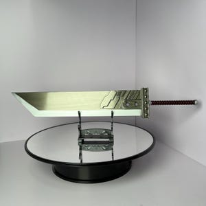 Könnte beinhalten: Eine große, silberfarbene Buster Sword-Replik mit detailliertem Knauf und burgunderfarbenem Griff. Die Klinge ist breit und flach, mit poliertem Finish. Das Schwert wird auf einem schwarzen Ständer mit reflektierender Oberfläche ausgestellt.