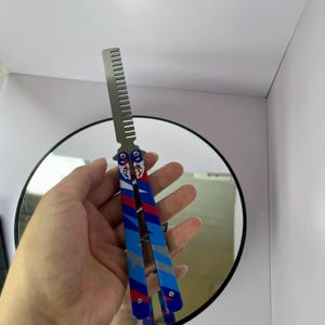 K&ouml;nnte beinhalten: Ein Butterfly-Kamm mit einer silbernen Kammklinge und einem Griff mit einem rot-wei&szlig;-blauen Design mit Stern- und Streifenmustern. Der Kamm wird vor einem runden Spiegel gehalten.