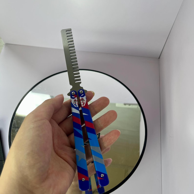 K&ouml;nnte beinhalten: Ein Butterfly-Kamm mit einer silbernen Kammklinge und einem Griff mit einem rot-wei&szlig;-blauen Design mit Stern- und Streifenmustern. Der Kamm wird vor einem runden Spiegel gehalten.