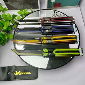 May include: A collection of colorful butterfly knives with black blades and handles in red, blue, yellow, and green. The knives are displayed on a reflective surface. A black leather case with the text "UM MODEL" is in the foreground.
