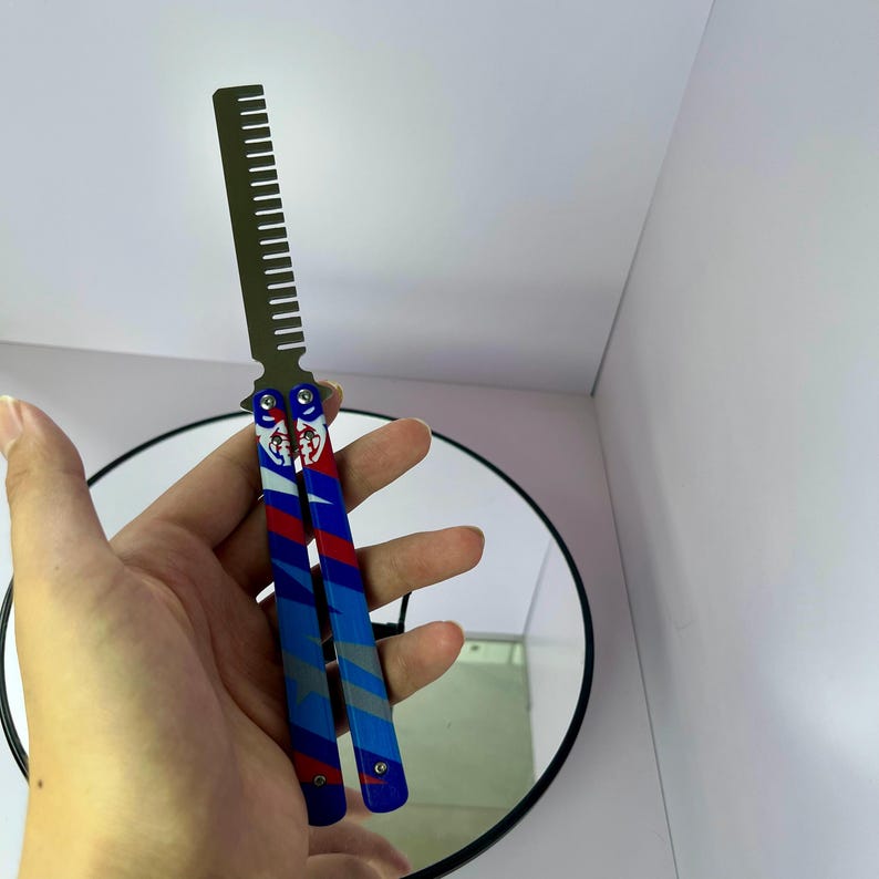 K&ouml;nnte beinhalten: Ein Kamm mit einem Butterfly-Messer-Design. Der Griff hat ein blaues, rotes und graues Streifenmuster mit einem dekorativen Emblem. Die Z&auml;hne des Kamms sind dunkelgrau, und der Griff ist ge&ouml;ffnet, um das Design zu zeigen.