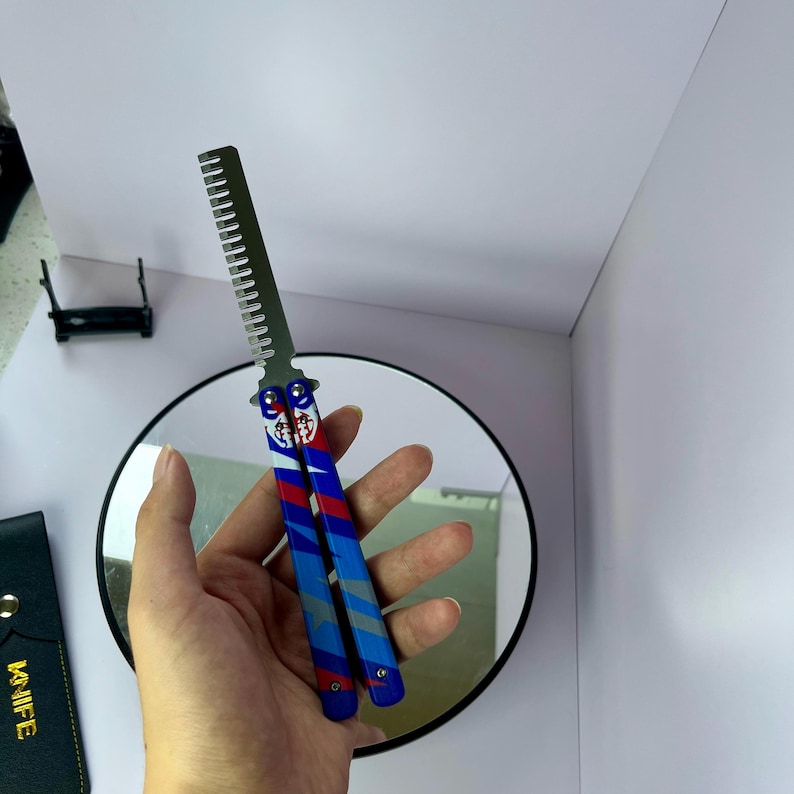 K&ouml;nnte beinhalten: Ein Butterflymesser mit Kammklinge. Der Griff ist blau, rot und wei&szlig; mit einem dekorativen Design. Das Messer wird vor einem runden Spiegel gehalten.