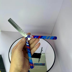 K&ouml;nnte beinhalten: Ein Butterflymesser mit Kammklinge. Der Griff hat ein rot-wei&szlig;-blaues Design. Der Kamm ist aus silberfarbenem Metall. Das Messer ist ge&ouml;ffnet und zeigt den Kamm. Hintergrund ist eine wei&szlig;e Wand und ein runder Spiegel.